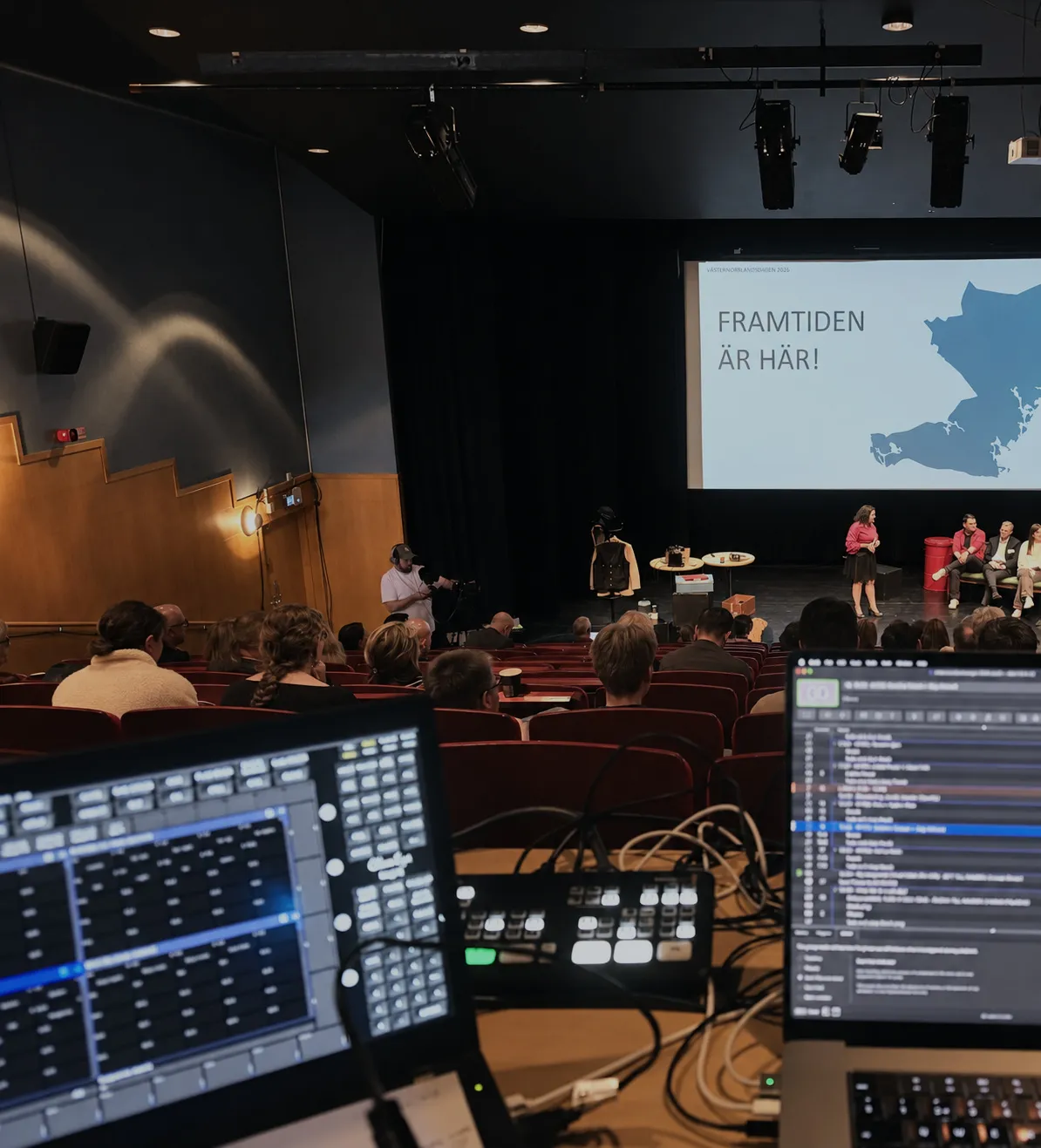 Bildmixer och skärmar vid regiplatsen, med kameraman och konferensscen i bakgrunden.